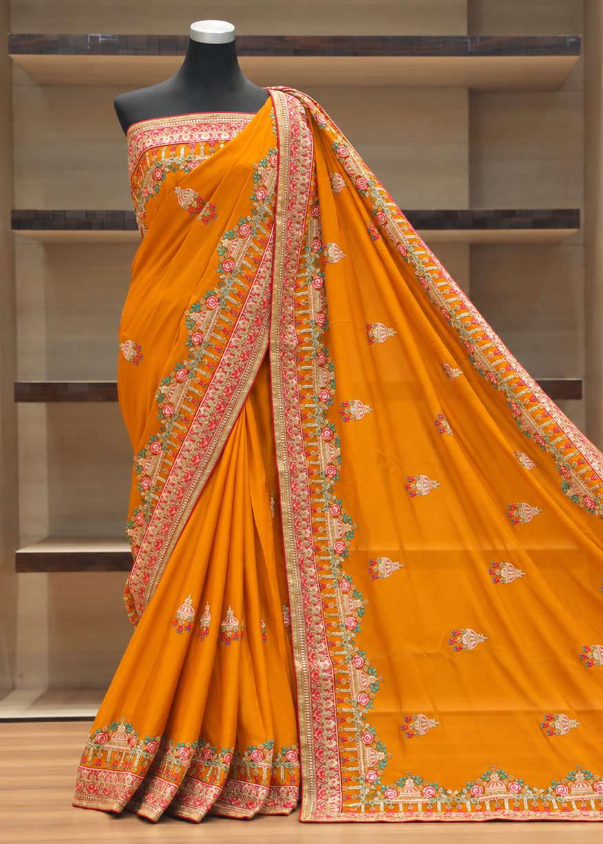 An orange saree with intricate multicolor borders and small floral motifs, displayed on a black mannequin torso in front of wooden shelving.