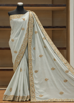 A light sage green saree with intricate gold and silver embroidery, displayed on a black mannequin torso against a wooden shelving background.