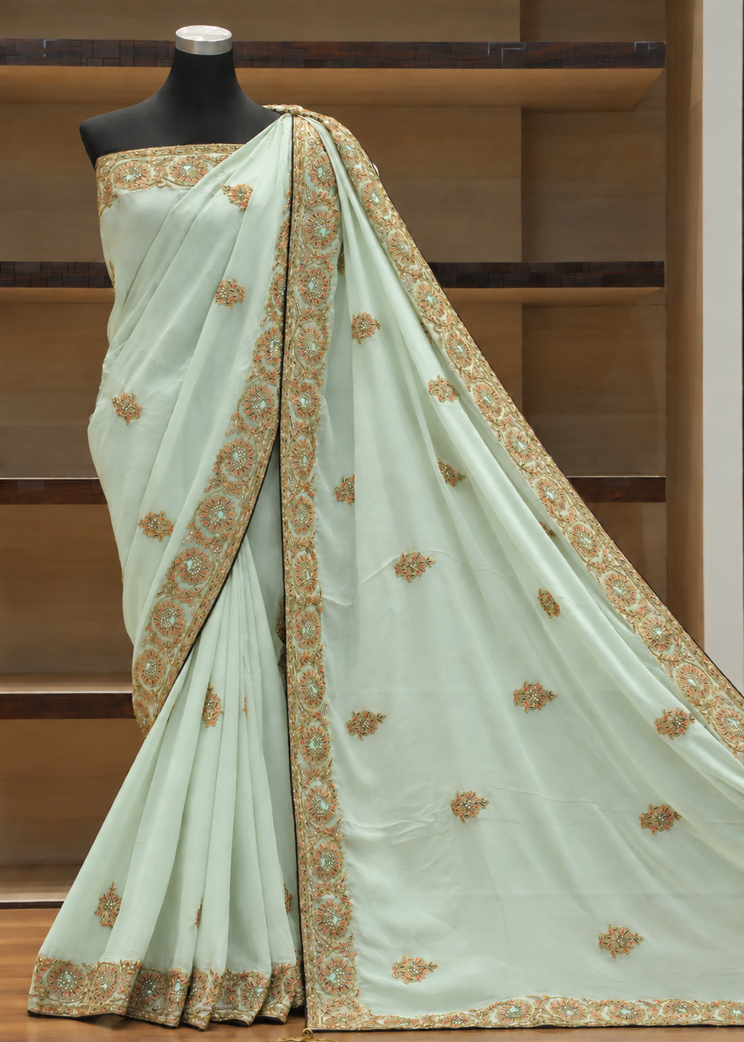 A pale mint green saree with intricate gold and silver embroidery, draped on a black mannequin torso against a wooden shelving background.