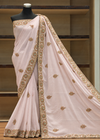 A light pink saree with intricate gold and green floral embroidery, draped on a black mannequin torso against a wooden shelf background.