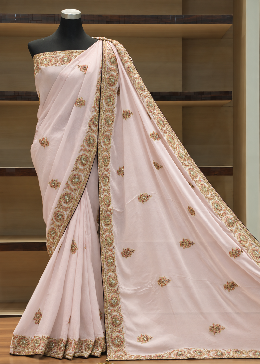 A light pink saree with intricate gold and green floral embroidery, draped on a black mannequin torso against a wooden shelf background.