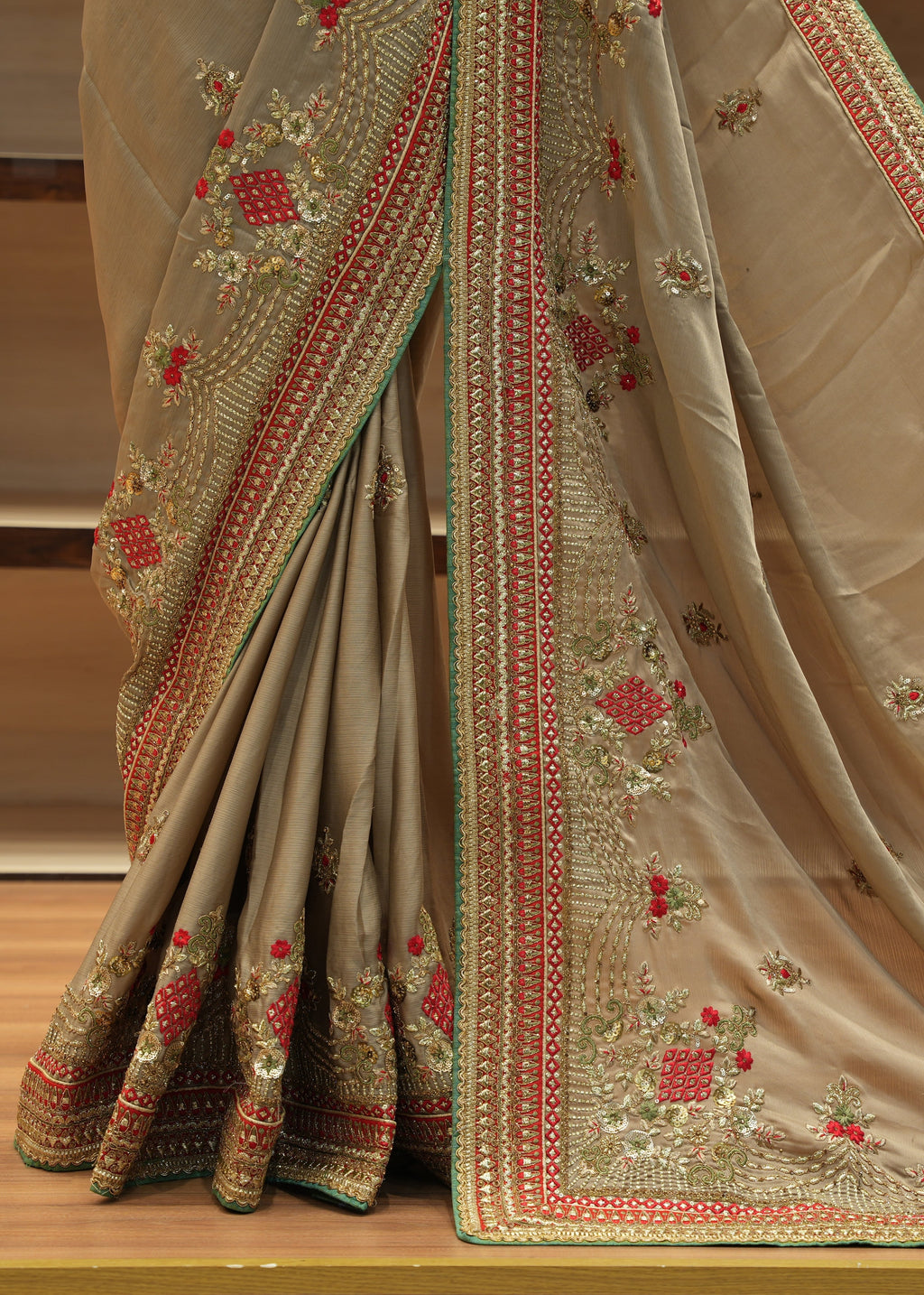 Beige saree with intricate red and gold embroidery on a wooden floor.
