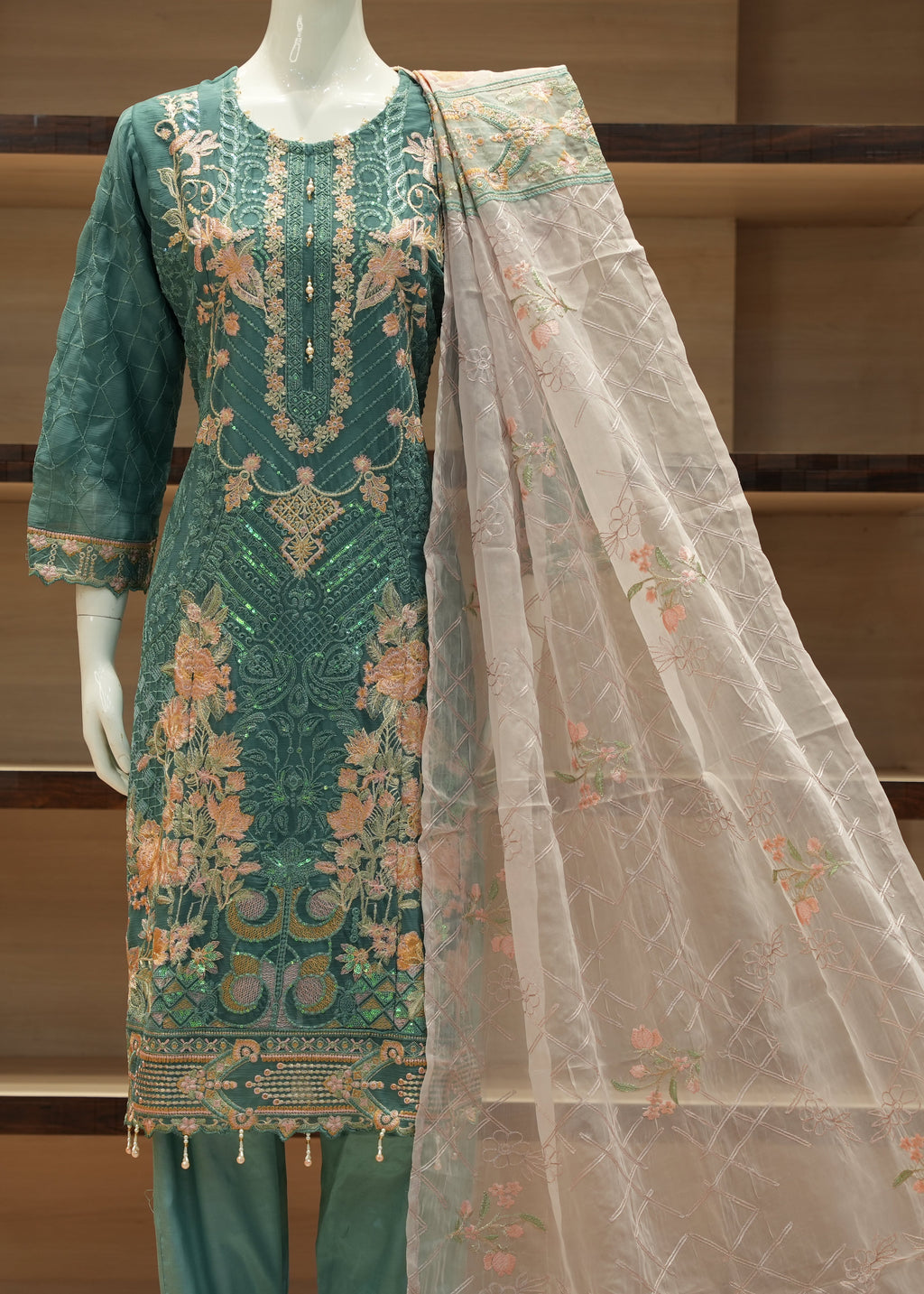 Green embroidered outfit with a sheer dupatta on a mannequin against a wooden shelf background