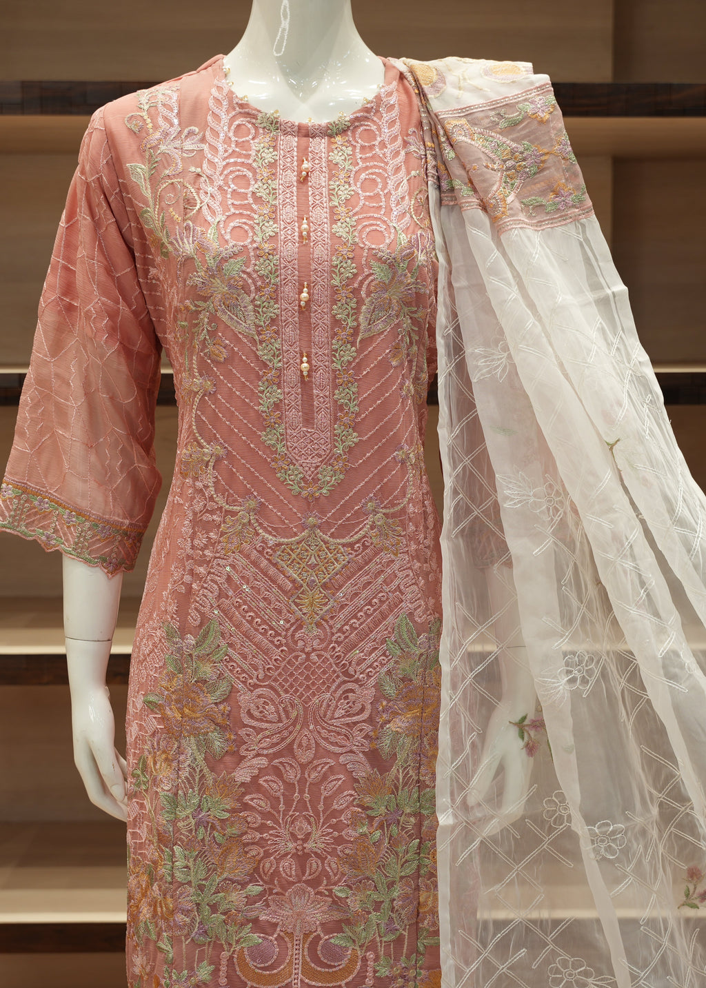 Pink embroidered dress with a white dupatta on a mannequin against a wooden shelf background