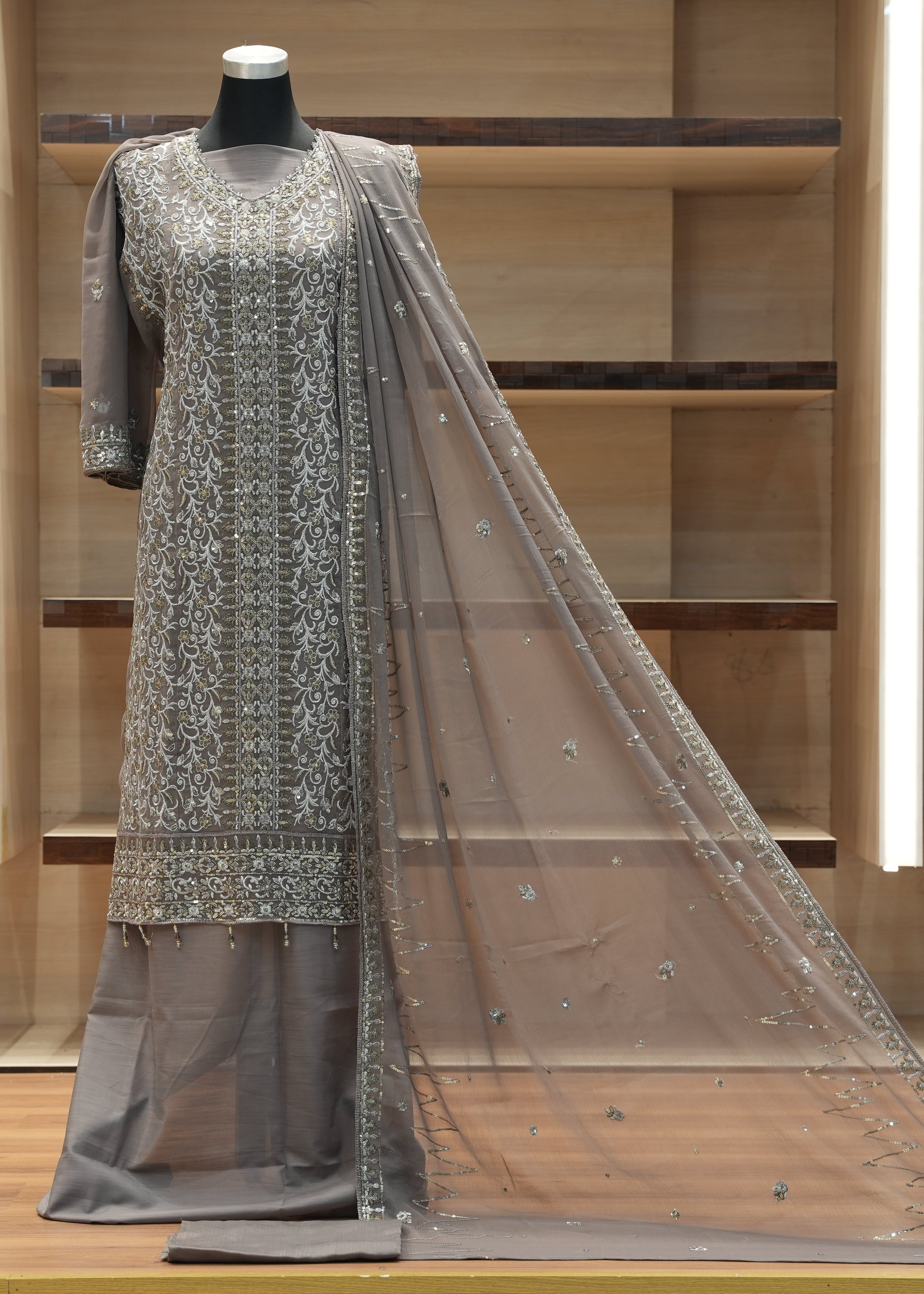 Gray embroidered traditional outfit with a sheer dupatta on a mannequin against a wooden shelf background.