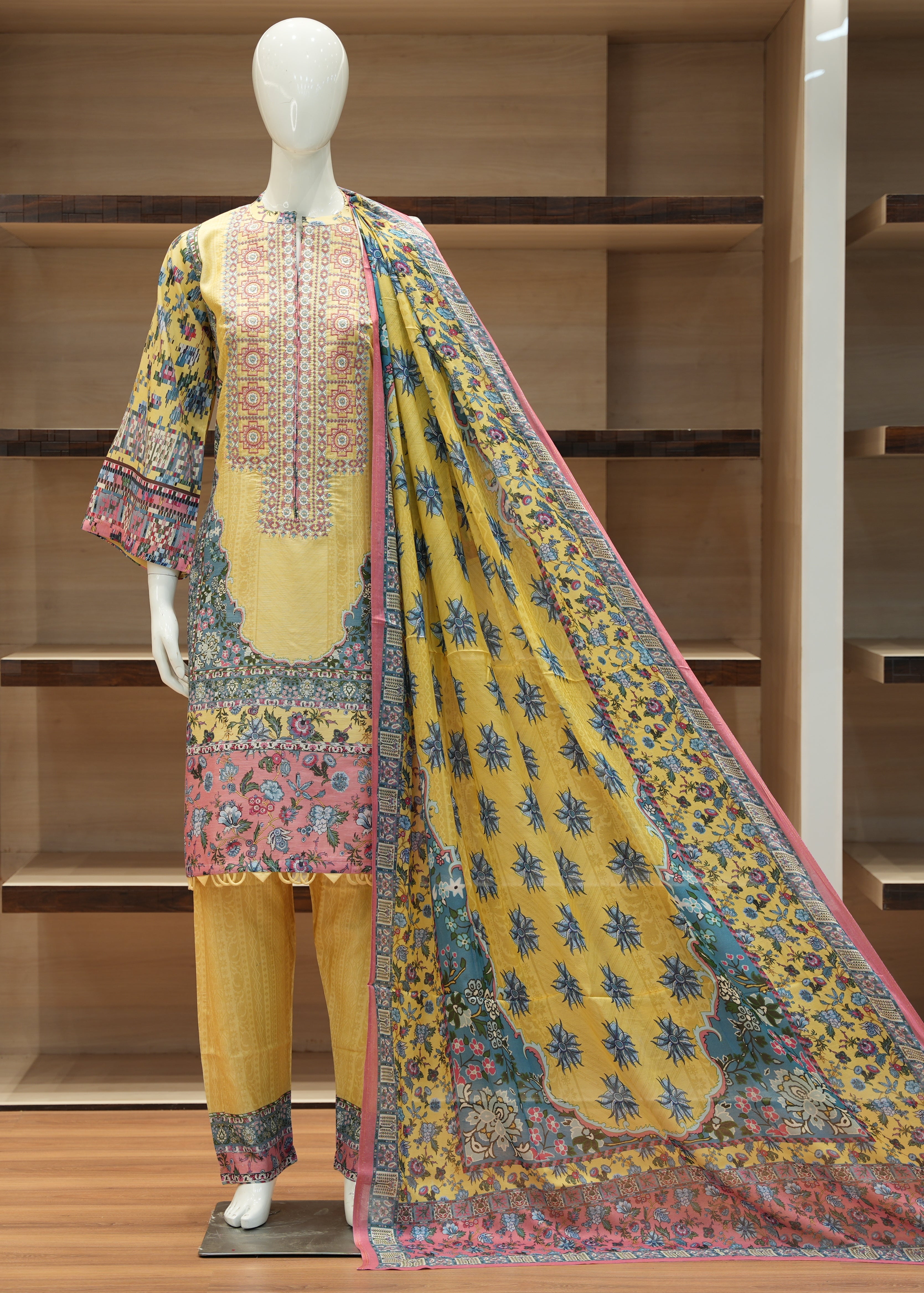 Mannequin wearing a traditional outfit with a yellow dress, multicolored dupatta, and embroidered top against a wooden background.