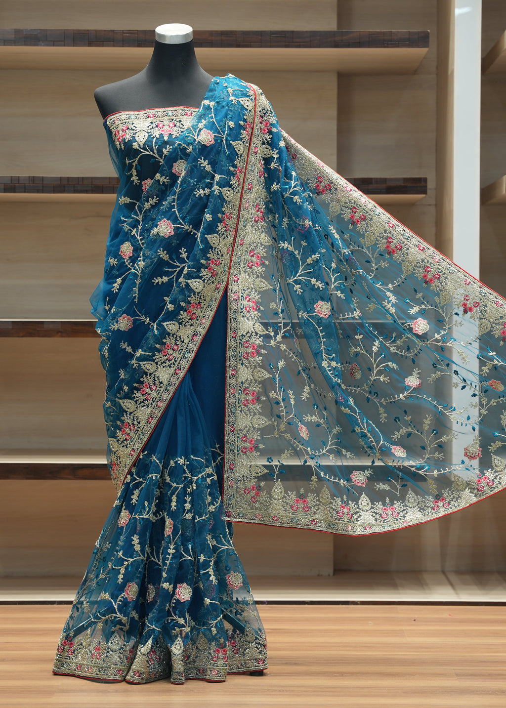 A teal blue embroidered saree displayed on a black mannequin in front of wooden shelving. The saree features intricate floral embroidery in pink, green, and gold, with a matching sheer dupatta draped over the shoulder.
