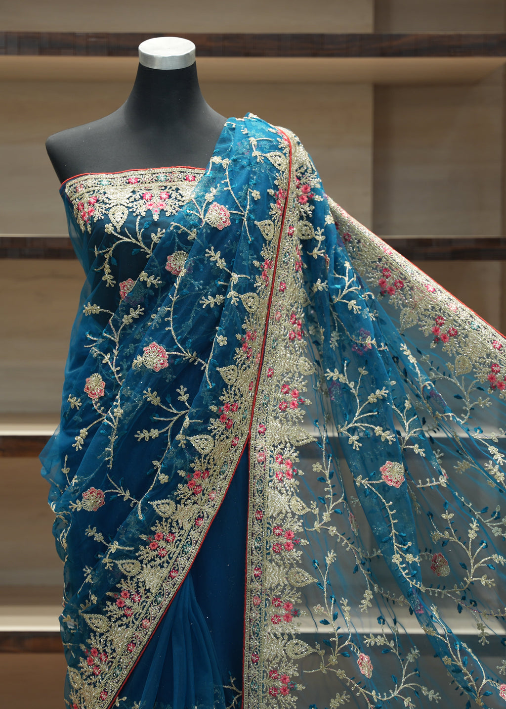 A blue embroidered saree displayed on a black mannequin torso, with the matching dupatta draped over the shoulder. The fabric features intricate floral embroidery in beige, pink, and red on a sheer mesh base, set against a wooden shelving background.