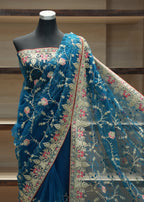 A blue embroidered saree displayed on a black mannequin torso, with the matching dupatta draped over the shoulder. The fabric features intricate floral embroidery in beige, pink, and red on a sheer mesh base, set against a wooden shelving background.