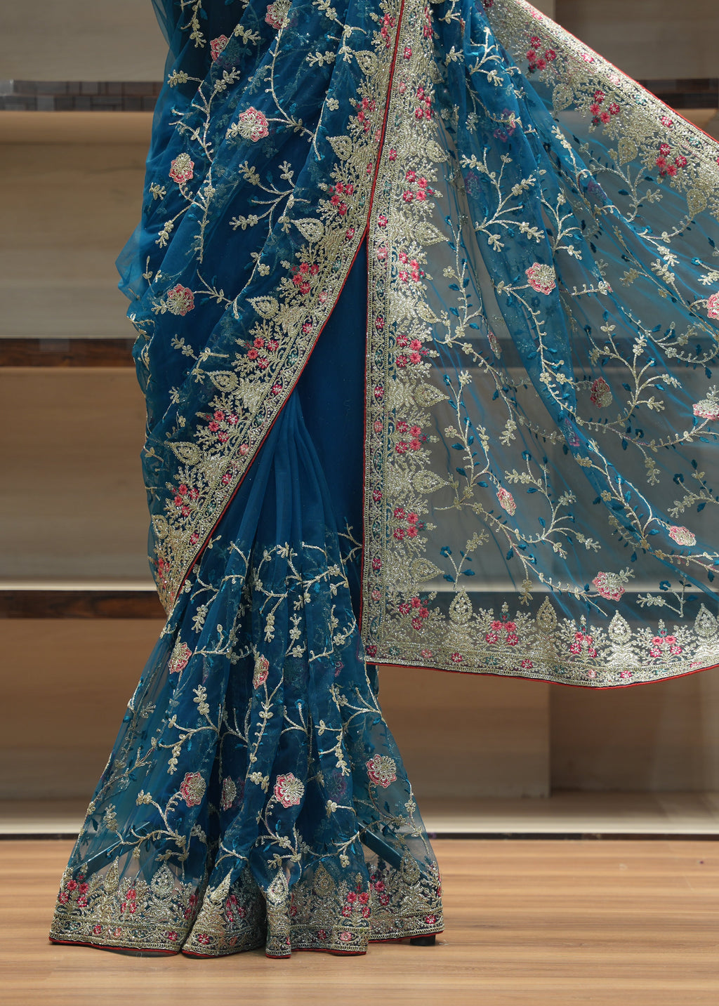 A teal blue saree with intricate floral embroidery and a matching sheer border, draped on a model in front of a wooden shelving unit.