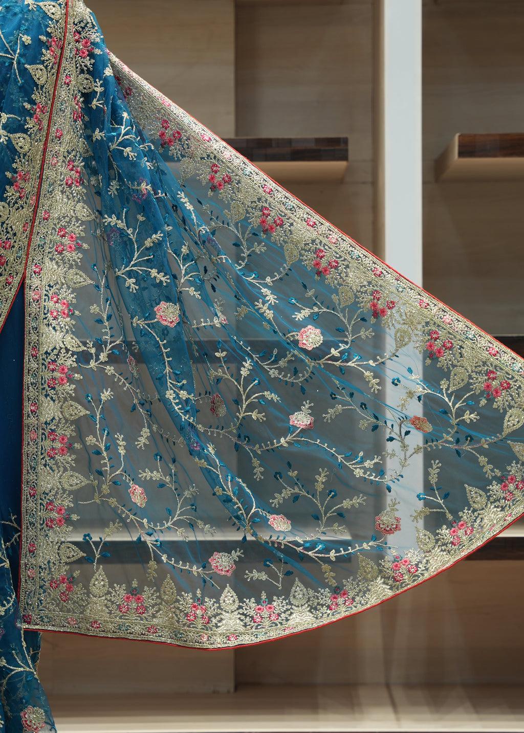 A teal blue embroidered saree drape or dupatta with floral vine motifs in gold, pink, and blue, shown against a wooden shelving background.