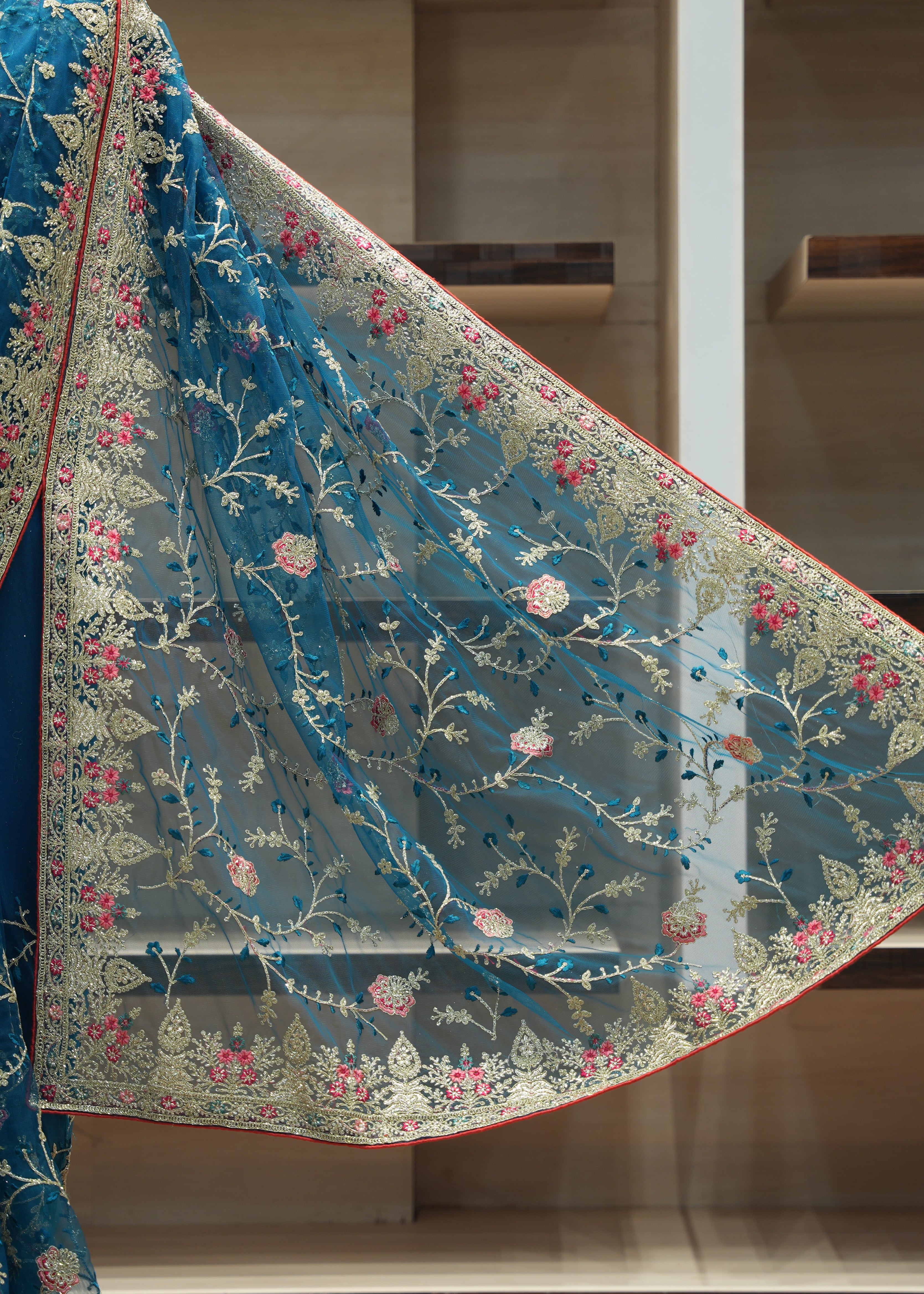 A teal blue embroidered saree drape or dupatta with floral vine motifs in gold, pink, and blue, shown against a wooden shelving background.