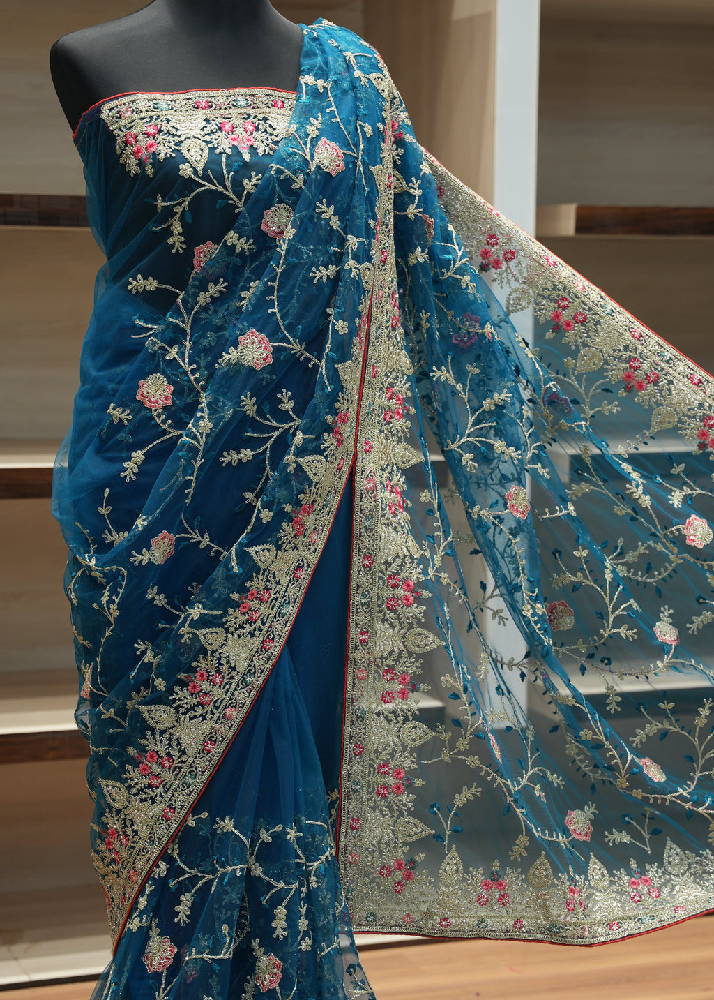 A teal blue embroidered saree draped on a black mannequin, featuring intricate floral vine embroidery in gold, pink, and white, with a matching sheer dupatta draped over the shoulder.
