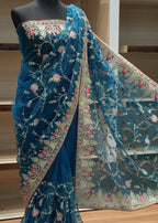A teal blue embroidered saree draped on a black mannequin, featuring intricate floral vine embroidery in gold, pink, and white, with a matching sheer dupatta draped over the shoulder.