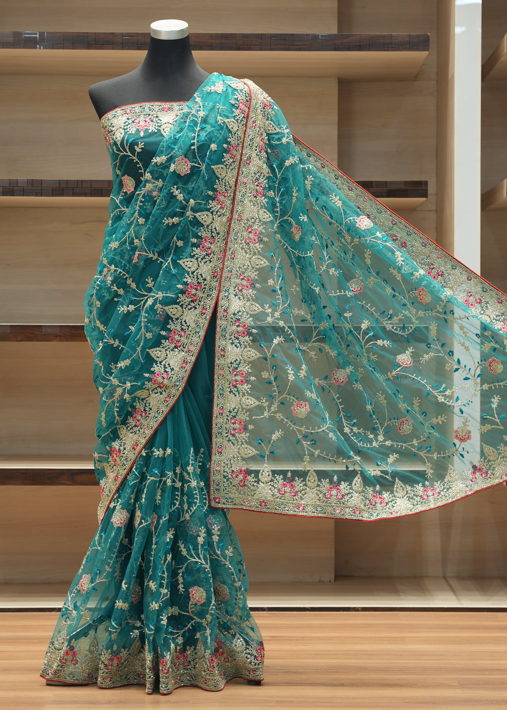 A teal green embroidered saree displayed on a black mannequin torso in front of wooden shelving. The saree features intricate floral embroidery in pink, gold, and white, with a matching sheer dupatta draped to the side.