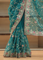 A teal green embroidered saree with pink floral motifs and gold zari detailing, draped to show the flared skirt and matching sheer dupatta.