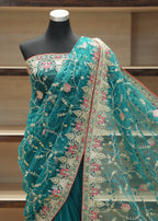 A teal green embroidered saree draped on a black mannequin torso, featuring intricate floral embroidery in pink, gold, and white, with a red border, set against a wooden shelving background.