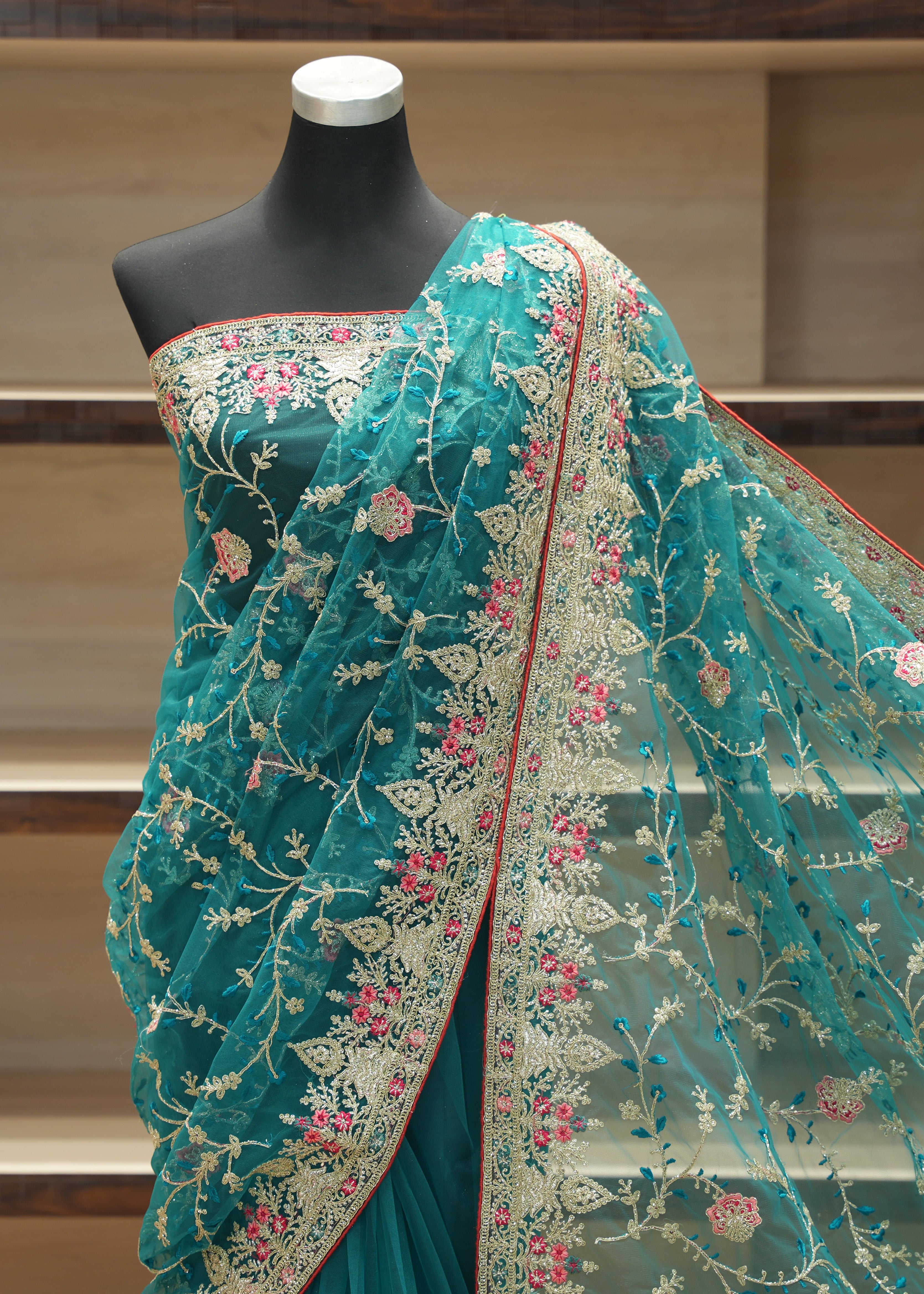 A teal green embroidered saree draped on a black mannequin torso, featuring intricate floral embroidery in pink, gold, and white, with a red border, set against a wooden shelving background.