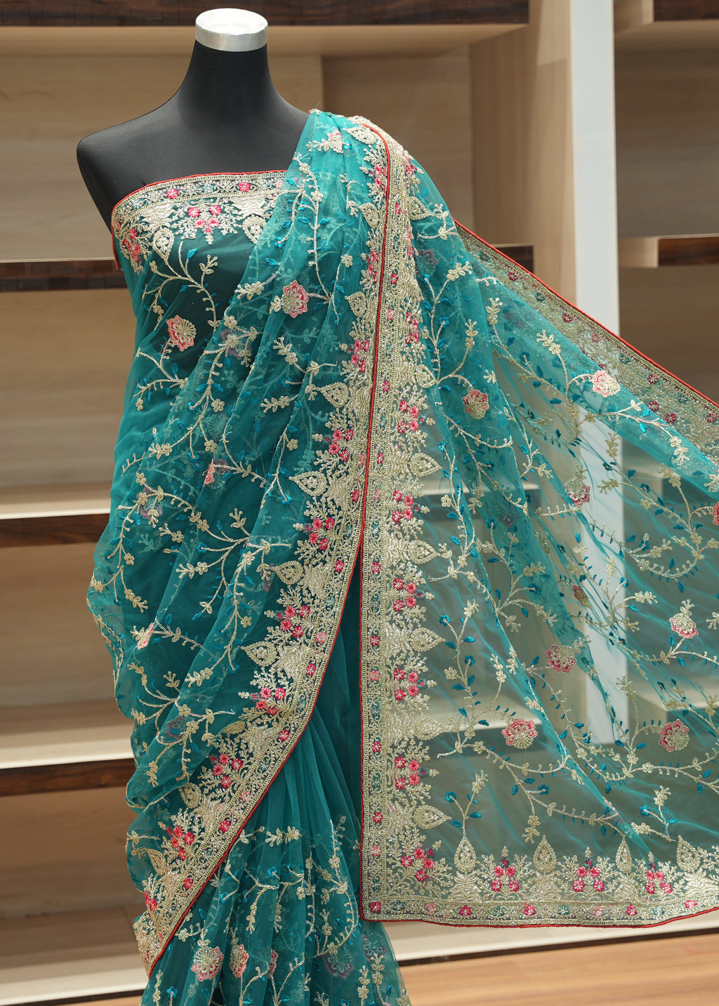 A teal green saree with intricate gold floral embroidery and pink floral accents, draped on a black mannequin torso. The matching sheer dupatta is spread out to the right, showcasing the detailed gold and pink embroidery.