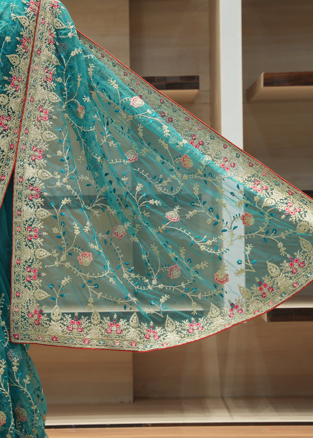 A teal green embroidered saree with a matching sheer dupatta draped over a mannequin, featuring intricate floral and vine patterns in gold, pink, and blue on a light beige base.