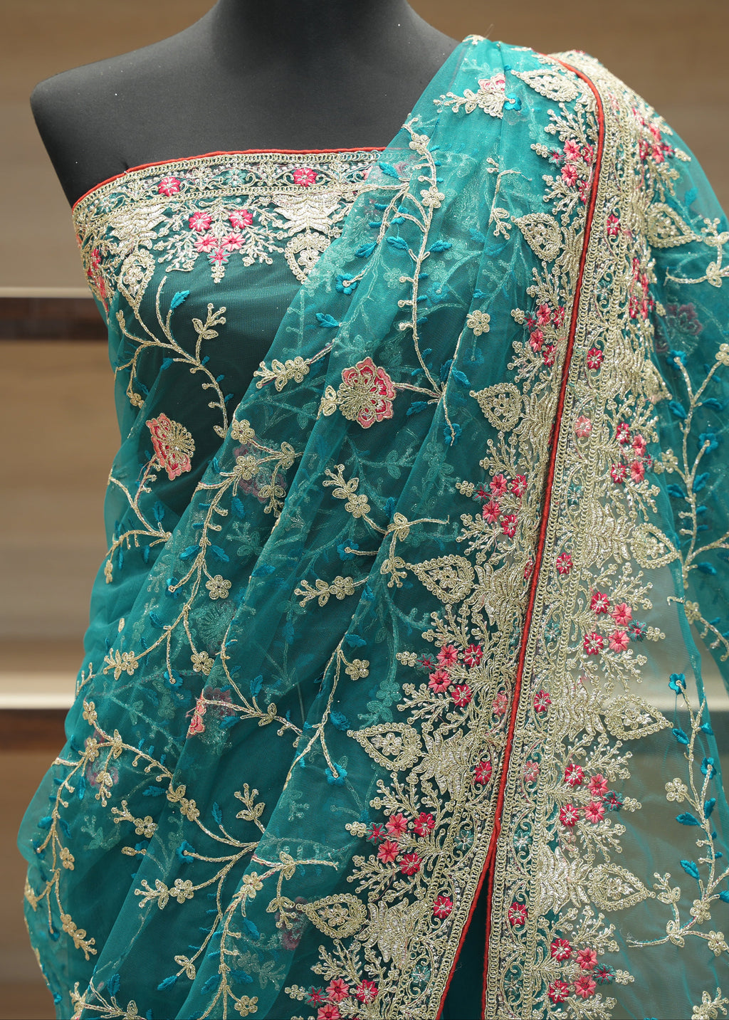A teal green embroidered saree draped on a black mannequin torso, featuring intricate gold floral embroidery, pink floral accents, and a red border, with a matching embroidered blouse.