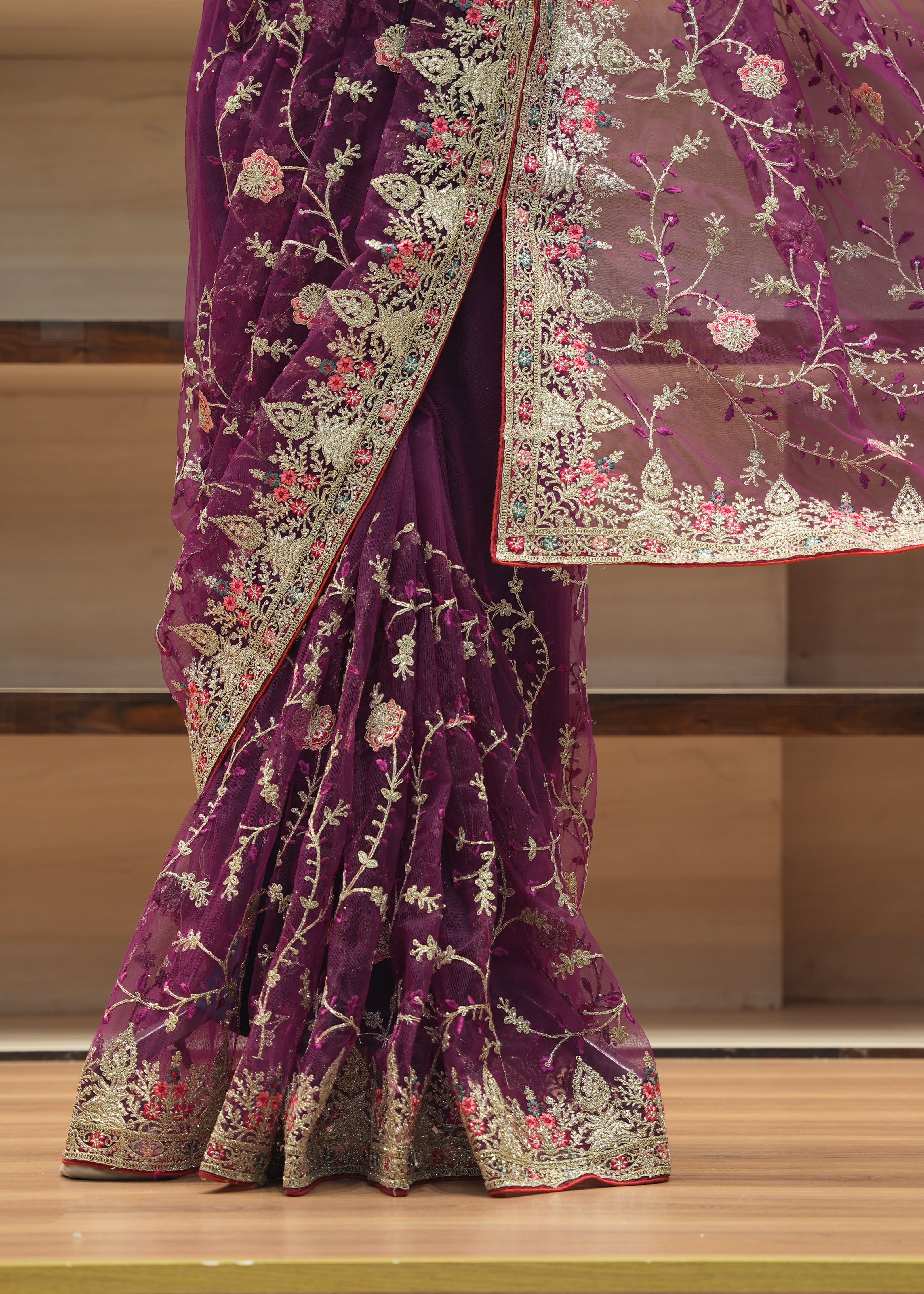 A close-up of a purple saree draped on a model, featuring intricate silver and gold floral embroidery and a matching sheer dupatta with floral patterns, set against a wooden shelf background.