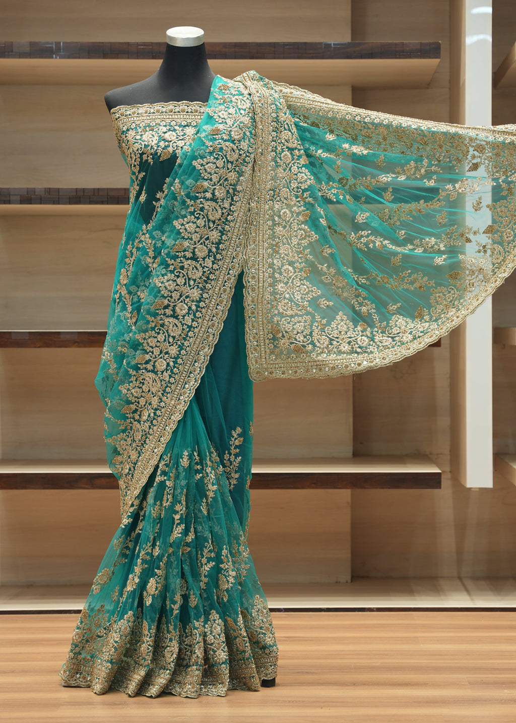 A teal green saree with intricate gold floral embroidery displayed on a black mannequin torso in front of wooden shelving.