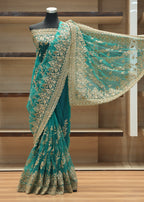 A teal green saree with intricate gold floral embroidery displayed on a black mannequin torso in front of wooden shelving.