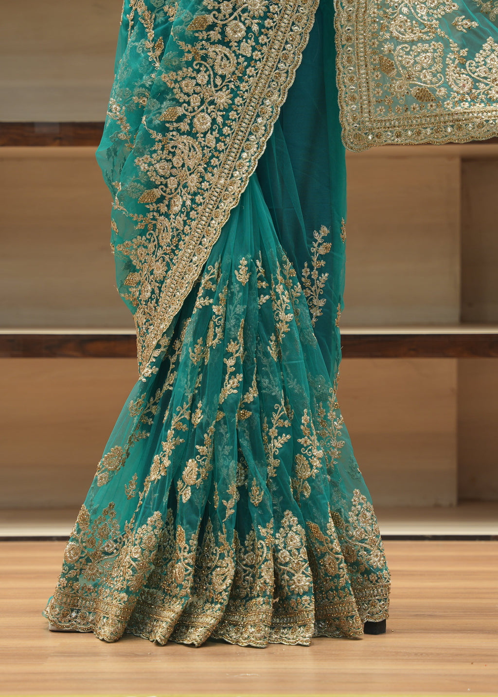 A teal green saree with intricate gold floral embroidery and a wide gold border, draped on a wooden floor in front of wooden shelves.