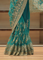 A teal green saree with intricate gold floral embroidery and a wide gold border, draped on a wooden floor in front of wooden shelves.