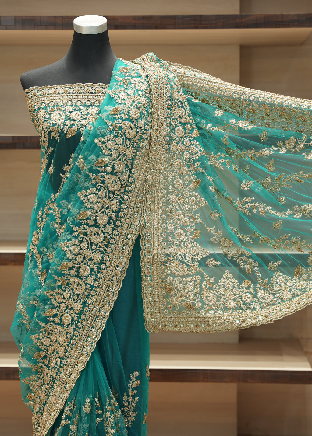 A teal green saree with intricate gold floral embroidery and a matching sheer dupatta draped on a black mannequin, set against a wooden shelving background.