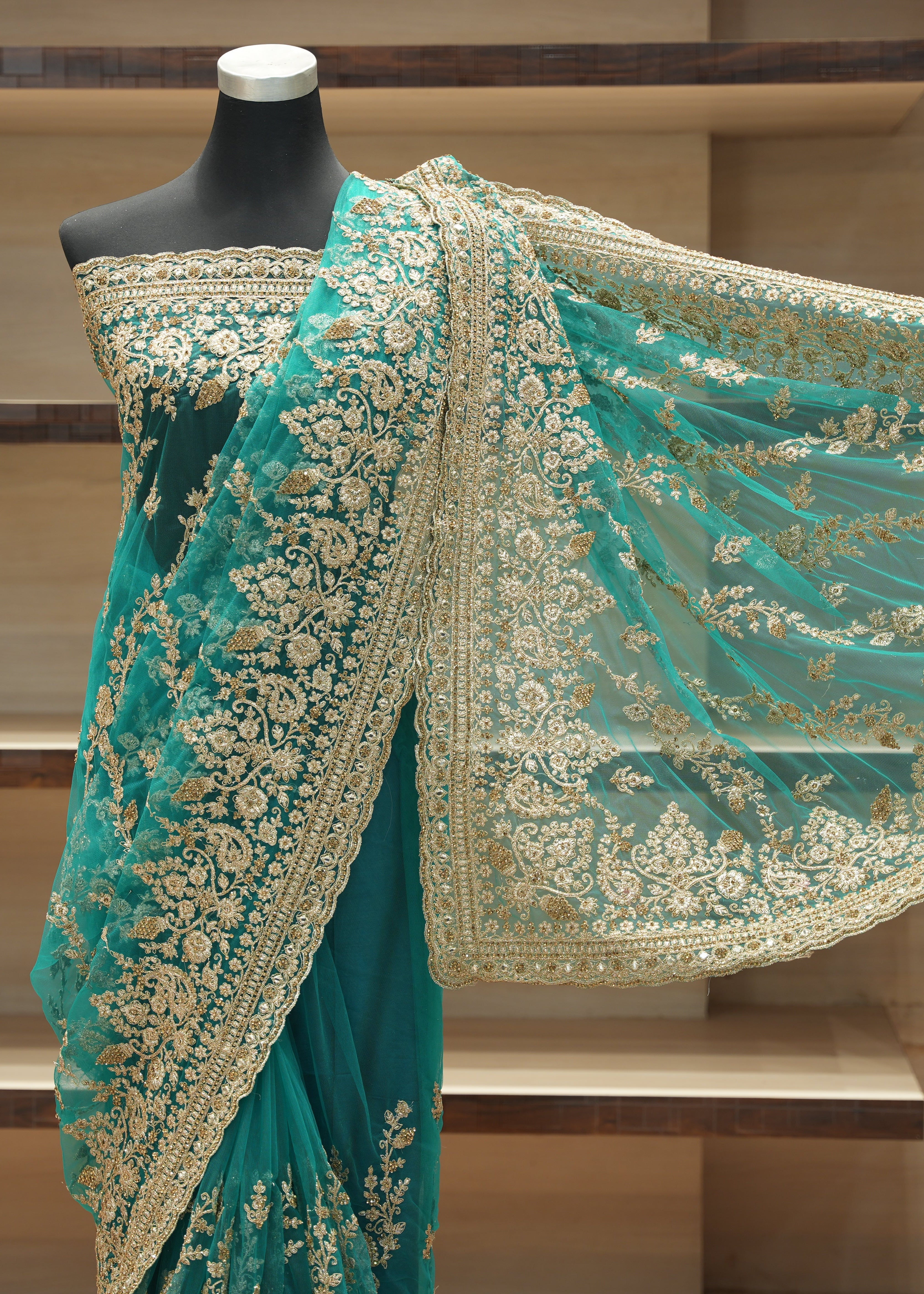 A teal green saree with intricate gold floral embroidery and a matching sheer dupatta draped on a black mannequin, set against a wooden shelving background.