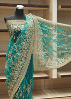 A teal green embroidered saree with gold floral patterns displayed on a black mannequin, with the matching sheer dupatta draped out to the side against a wooden shelving background.