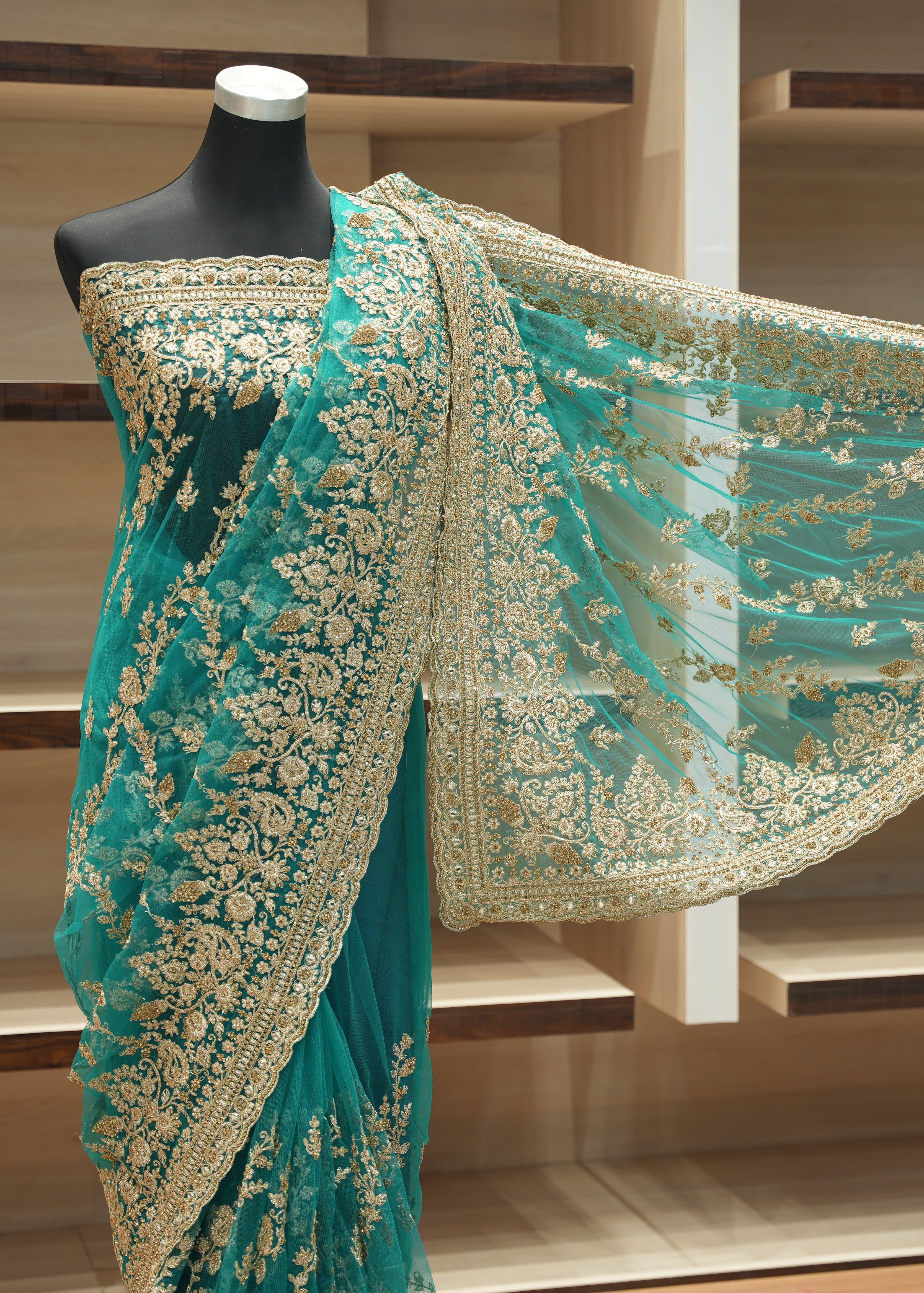 A teal green embroidered saree with gold floral patterns displayed on a black mannequin, with the matching sheer dupatta draped out to the side against a wooden shelving background.