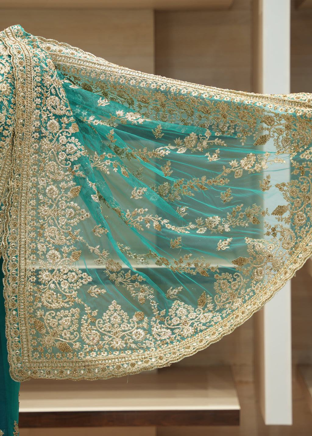 A close-up of a teal green embroidered fabric accessory, likely a dupatta or sleeve overlay, featuring intricate gold floral embroidery and a scalloped border, displayed against a blurred indoor background.
