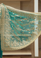 A close-up of a teal green embroidered fabric accessory, likely a dupatta or sleeve overlay, featuring intricate gold floral embroidery and a scalloped border, displayed against a blurred indoor background.