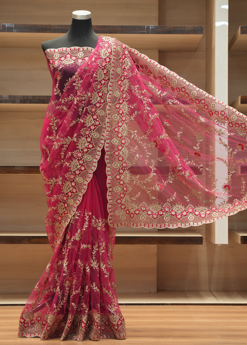 A bright pink georgette saree with intricate gold floral embroidery, displayed on a black mannequin torso in front of wooden shelving.
