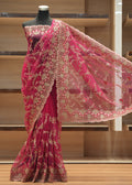 A bright pink georgette saree with intricate gold floral embroidery, displayed on a black mannequin torso in front of wooden shelving.