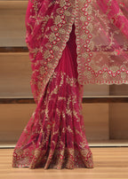 A close-up of a bright pink saree with intricate gold floral embroidery and a matching sheer pink dupatta draped over a wooden shelf.