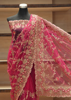 A bright pink georgette saree with intricate gold floral embroidery and a matching embroidered border, displayed on a black mannequin torso in front of wooden shelves.