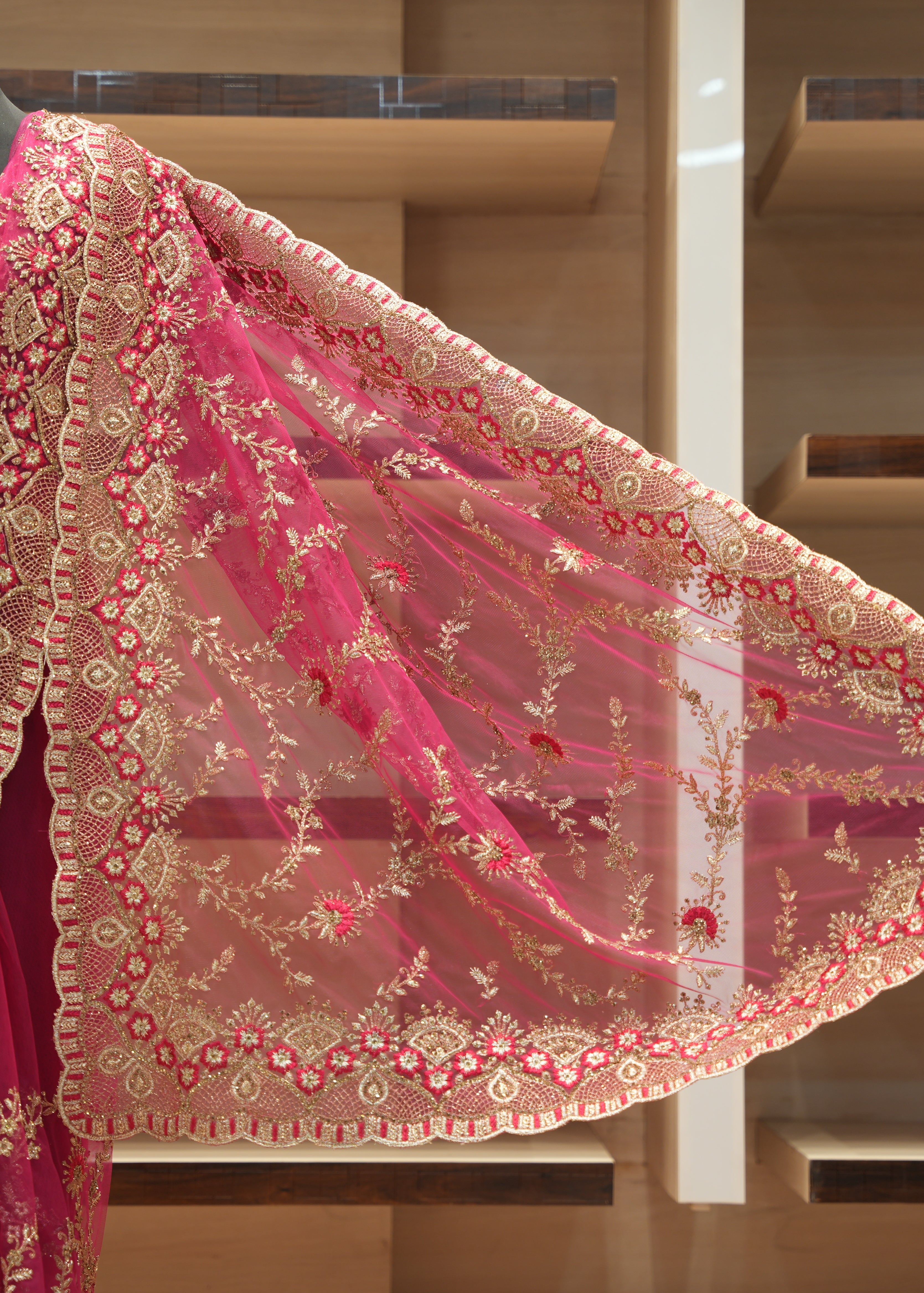 A close-up of a pink embroidered saree drape or dupatta with ornate gold floral and geometric patterns, displayed on a mannequin in front of wooden shelving.