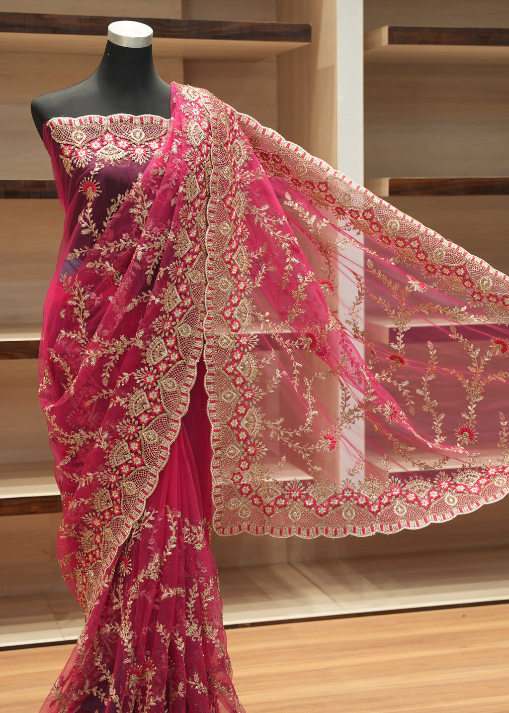 A pink and gold embroidered saree displayed on a black mannequin torso, with the fabric draped to show the border and pattern details against a wooden shelving background.