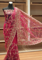 A pink and gold embroidered saree displayed on a black mannequin torso, with the fabric draped to show the border and pattern details against a wooden shelving background.