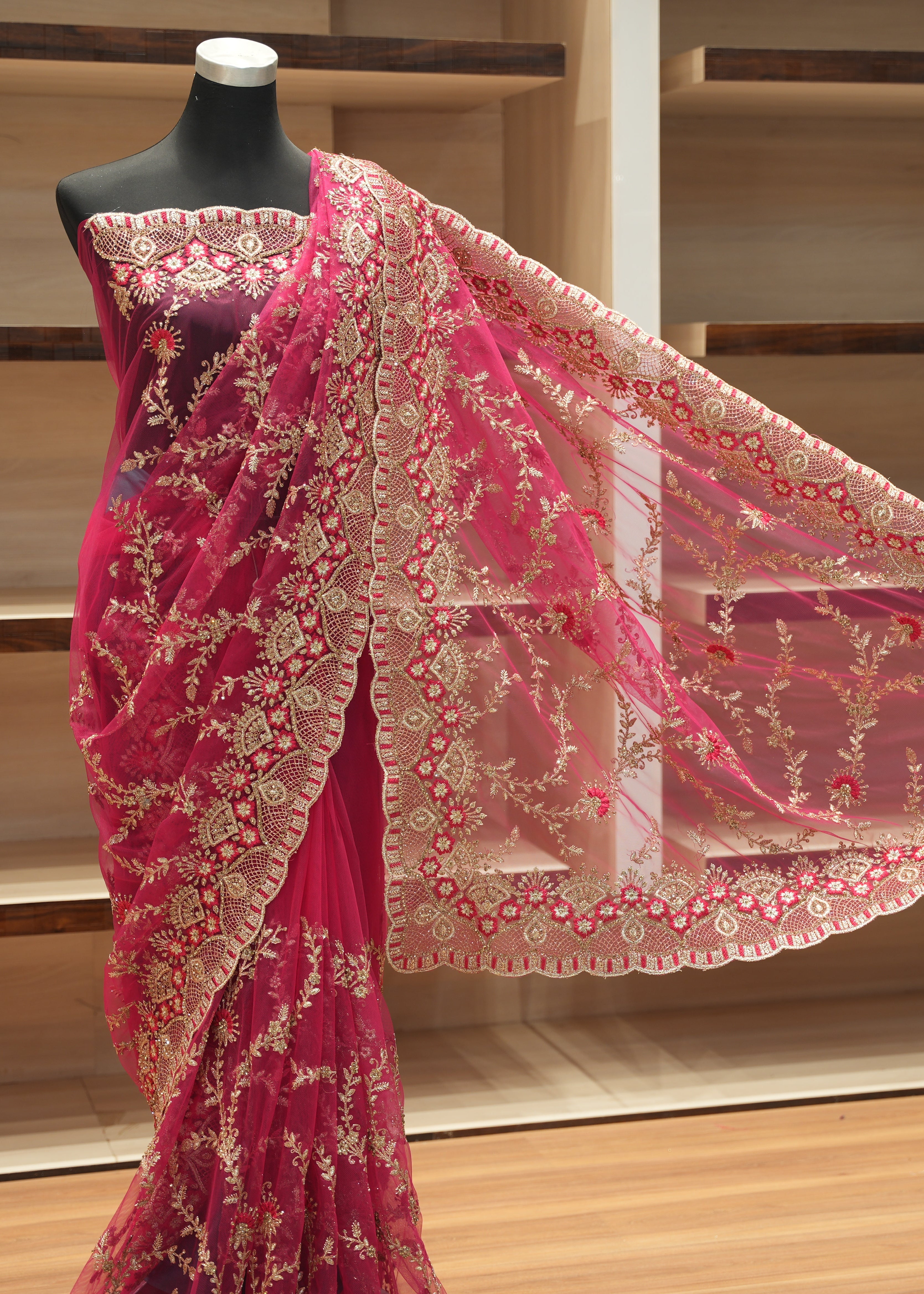 A pink and gold embroidered saree displayed on a black mannequin torso, with the fabric draped to show the border and pattern details against a wooden shelving background.