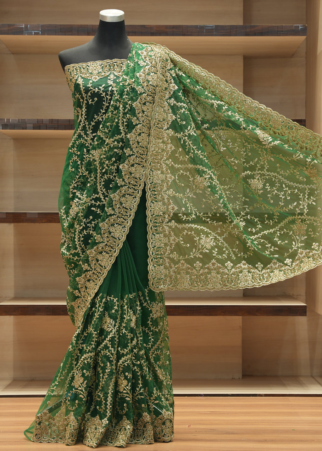 A dark green saree with intricate gold embroidery and a matching sheer dupatta draped on a black mannequin, set against a wooden shelving background.