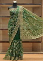 A dark green saree with intricate gold embroidery and a matching sheer dupatta draped on a black mannequin, set against a wooden shelving background.