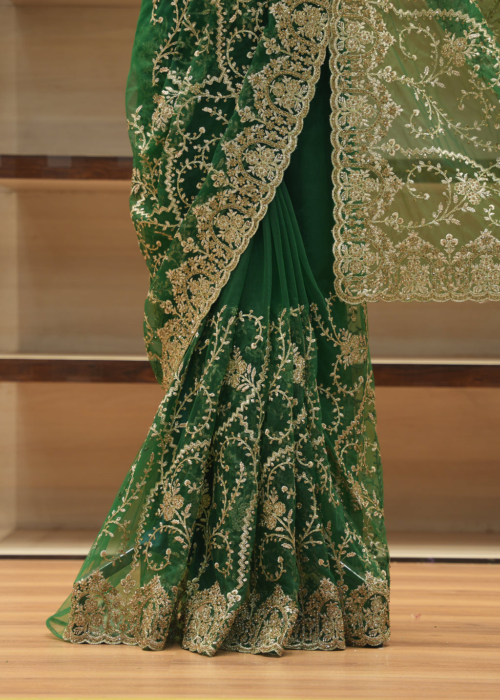 A close-up of a green saree with intricate gold floral embroidery and a matching sheer border, draped on a wooden floor in front of a wooden shelving unit.