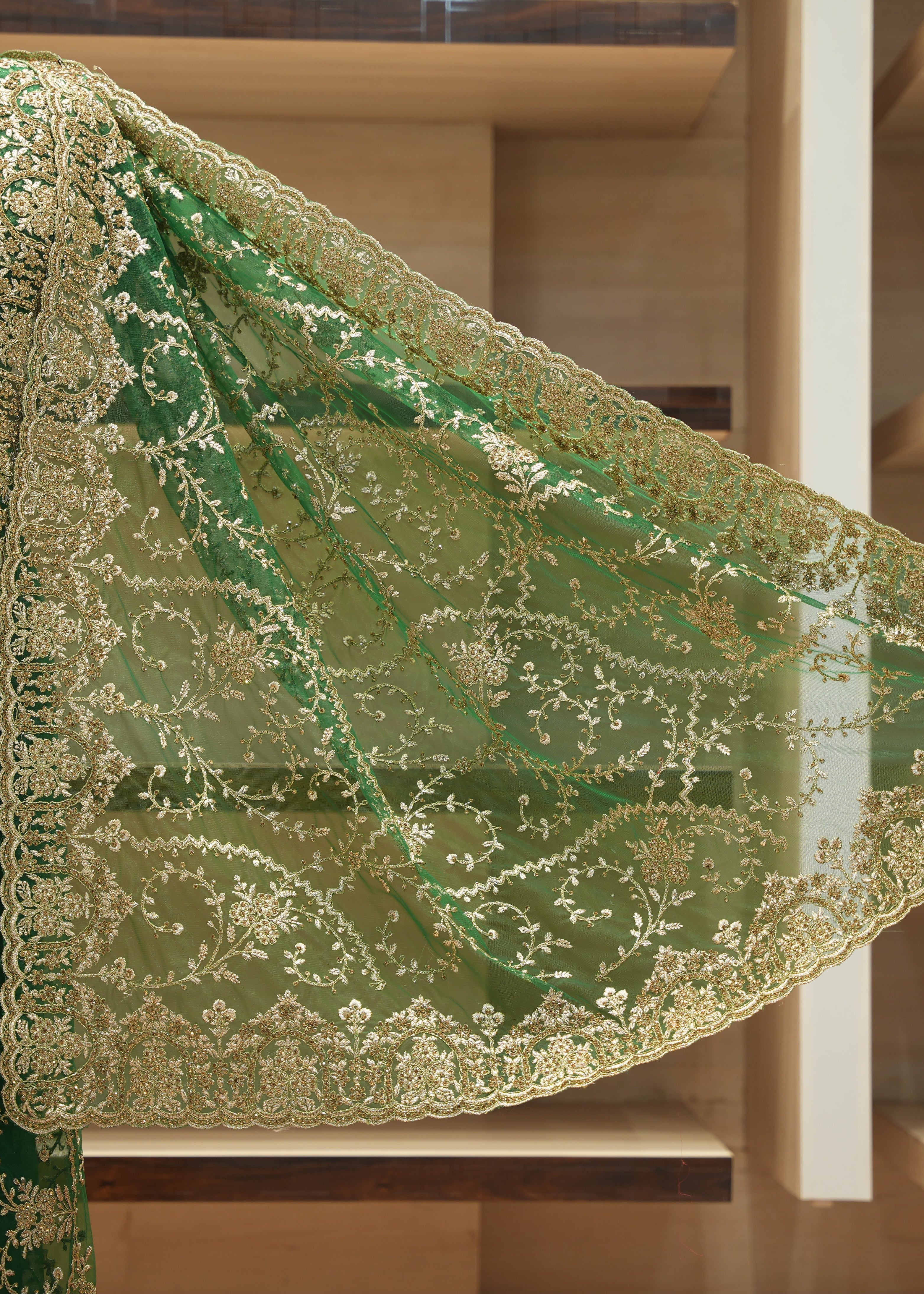A green embroidered fabric drape with ornate gold floral lace trim, displayed on a mannequin torso in front of wooden shelving.