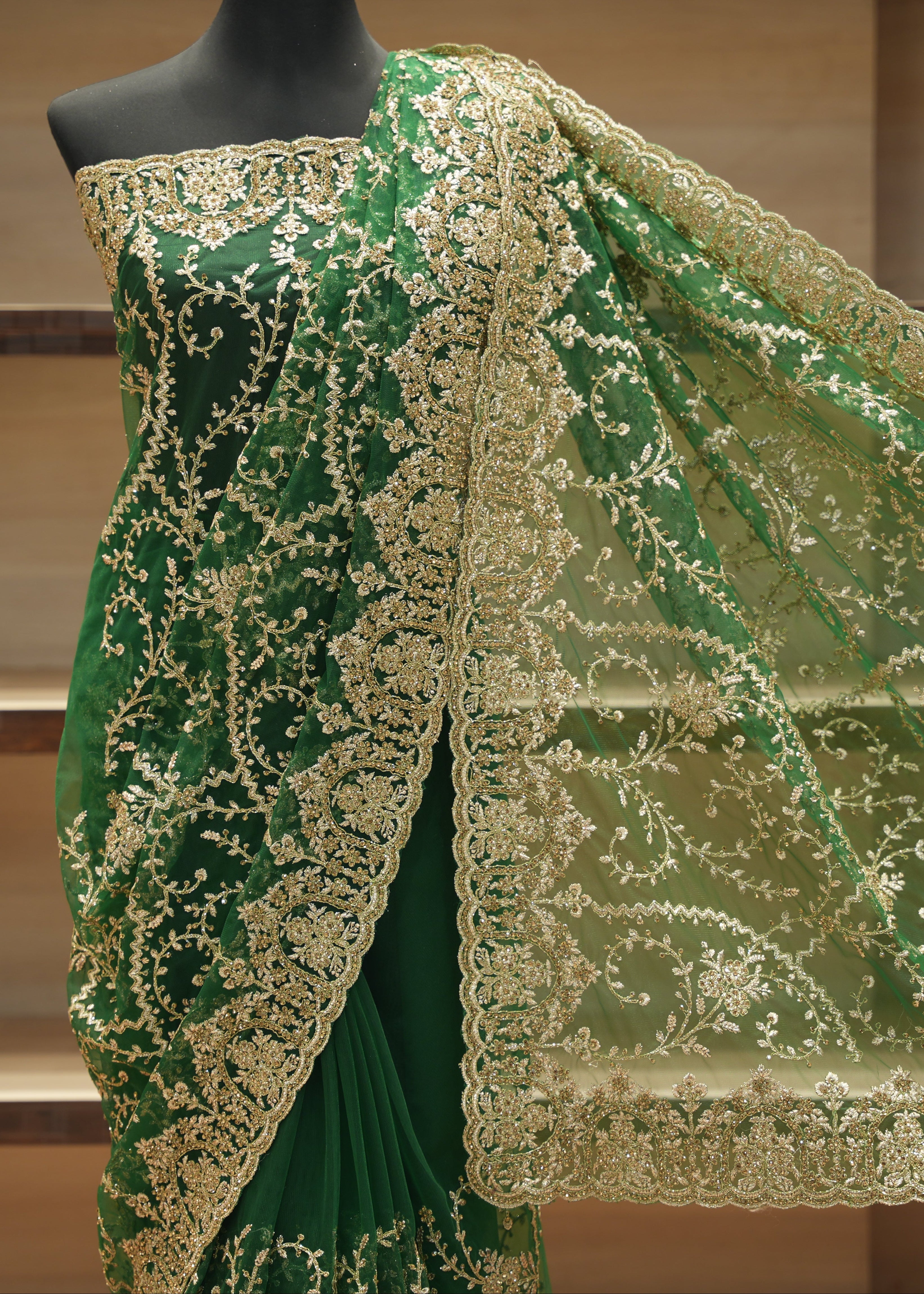 A green saree draped on a black mannequin, featuring intricate gold floral embroidery and a matching sheer dupatta with a wide gold border, set against a wooden shelving background.