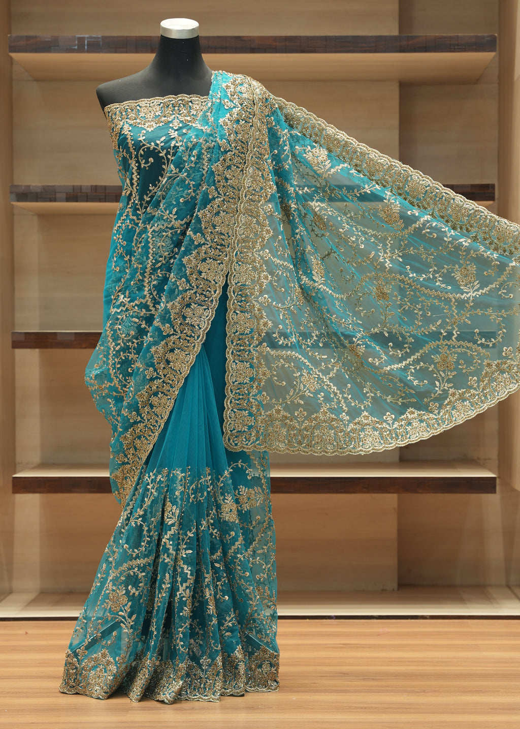 A teal blue saree with intricate gold floral embroidery and a wide matching border, displayed on a black mannequin in front of wooden shelving.
